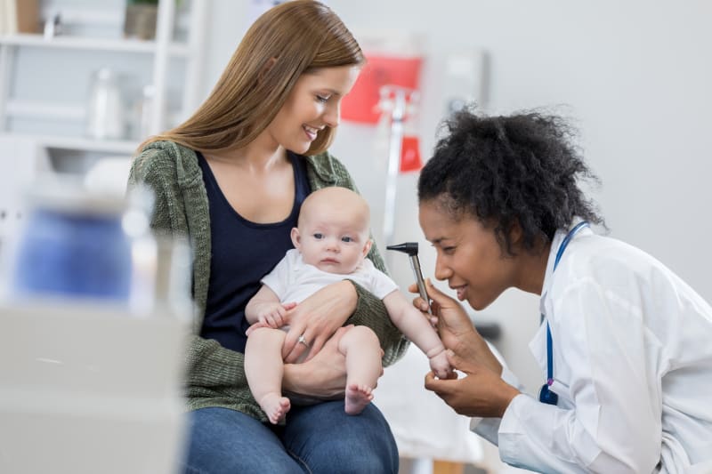 Mother holding young baby while female doctor examines baby's ear with otoscope.