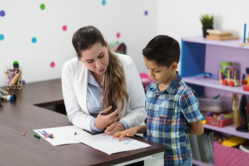 A latin female psychologist listening to a little boy in her office talking about his drawing.