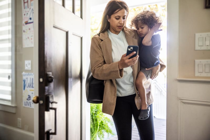 Mother walking through doorway dialing smartphone while holding toddler boy.